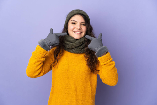Adolescente rusa chica con sombrero de invierno aislado sobre fondo púrpura dando un gesto pulgares hacia arriba - Foto, imagen
