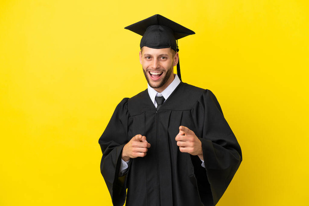 Fiatal egyetemi diplomás kaukázusi férfi elszigetelt sárga háttér mutatva az első és mosolygós - Fotó, kép