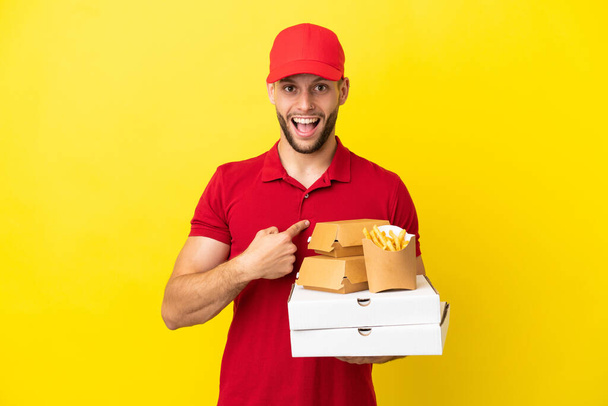 Доставщик пиццы подбирает коробки пиццы и бургеры на изолированном фоне с неожиданным выражением лица - Фото, изображение