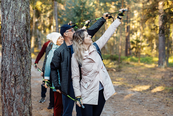 Group of people holding together Nordic trekking poles in the air on trek while hiking in the forest, nature. Exercises activity education of people of different ages, breathing and posture technic - Foto, Imagen