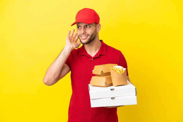 pizza delivery man picking up pizza boxes and burgers over isolated background listening to something by putting hand on the ear - Foto, Bild