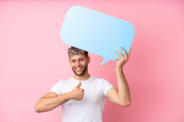 Young handsome caucasian man isolated on pink background holding an empty speech bubble with thumb up - 写真・画像