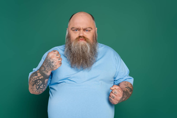 θυμωμένος υπέρβαρος άντρας με τατουάζ και γενειάδα που δείχνει σφιγμένες γροθιές απομονωμένες στο πράσινο - Φωτογραφία, εικόνα