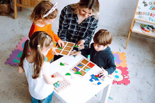 Kisgyerekek a pedagógus hajtogatás színes részleteket építész az asztalon a játszószobában felülnézetből. Érdekes játék az óvodások fejlődő logika és intelligencia. Kommunikáció a társakkal. - Fotó, kép