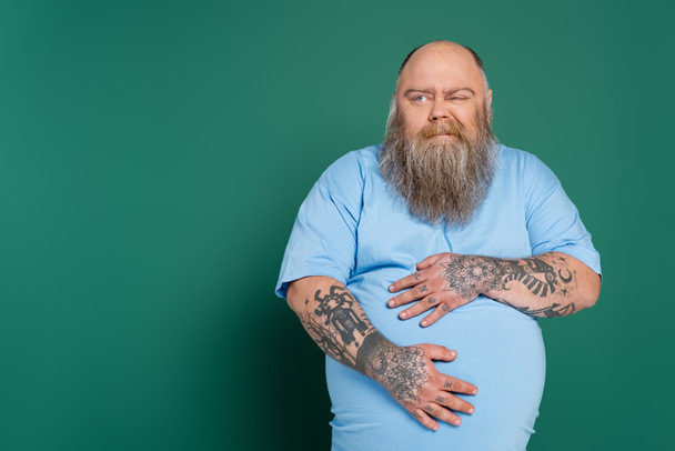 displeased overweight man hugging tummy while suffering from stomach pain isolated on green - Foto, imagen
