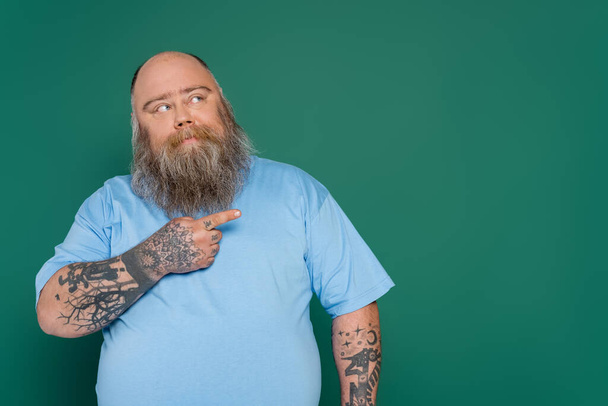 στοχαστικός υπέρβαρος άντρας με τατουάζ που δείχνει με το δάχτυλο και κοιτάζει μακριά απομονωμένος στο πράσινο - Φωτογραφία, εικόνα