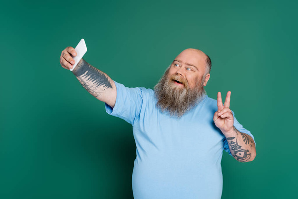 happy and bearded overweight man showing victory sign while taking selfie isolated on green - 写真・画像