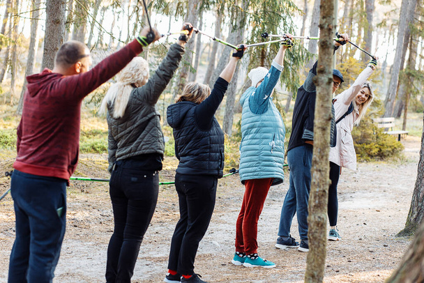 Senior people with female coach leader hold Nordic trekking sticks in air while hiking in the forest, nature. Exercises activity education, raised arms, prepare to hiking, breath and posture technic - Fotó, kép