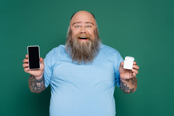cheerful plus size man with mobile phone and wireless earphones isolated on green - Foto, Imagen