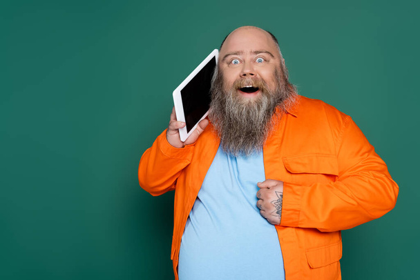 barbudo hombre asombrado con sobrepeso celebración tableta digital cerca de la cara aislada en verde - Foto, Imagen