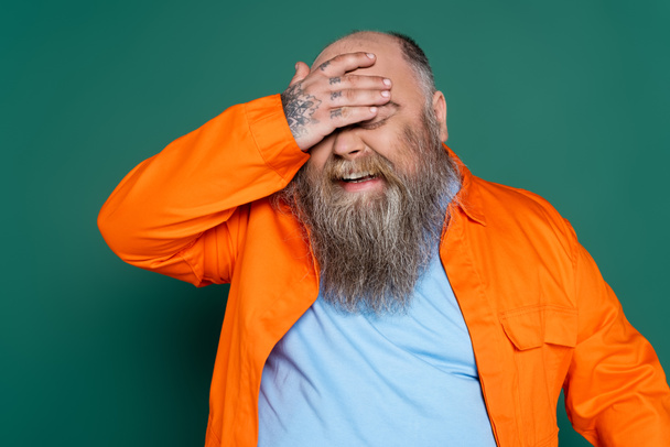 bearded overweight man laughing and covering eyes with hand isolated on green - Фото, зображення