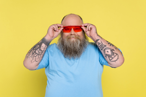 παχουλός και με τατουάζ άνθρωπος ρύθμιση μοντέρνα γυαλιά ηλίου, ενώ χαμογελά στην κάμερα απομονώνονται σε κίτρινο - Φωτογραφία, εικόνα