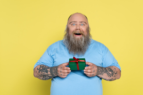 изумленный бородатый мужчина с избыточным весом держа зеленый подарочный коробок изолирован на желтый - Фото, изображение