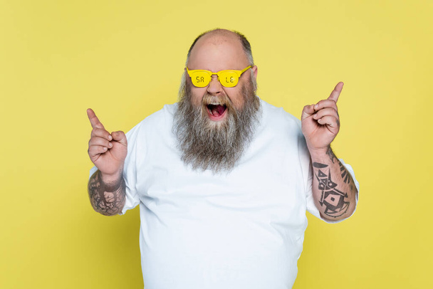 τολμηρός και γενειοφόρος υπέρβαρος άντρας σε γυαλιά με γράμματα πώλησης που δείχνουν με τα δάχτυλα απομονώνονται σε κίτρινο - Φωτογραφία, εικόνα