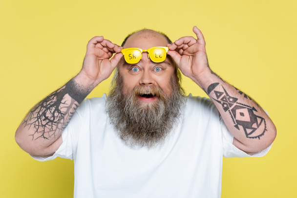 έκπληκτος τατουάζ συν μέγεθος άνθρωπος κρατώντας γυαλιά με την πώληση επιστολόχαρτα απομονώνονται σε κίτρινο - Φωτογραφία, εικόνα