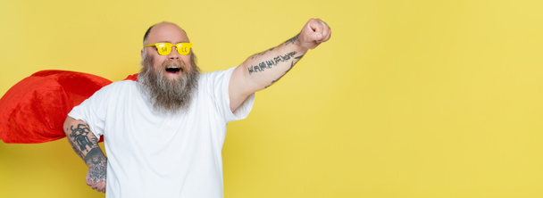 vzrušený nadváha muž v superhrdina plášť a brýle s prodejem písmo izolované na žluté, banner - Fotografie, Obrázek