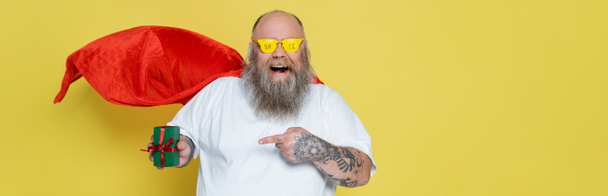 hombre excitado más tamaño en capa roja y gafas de sol con letras de la venta apuntando a la caja de regalo aislado en amarillo, bandera - Foto, imagen