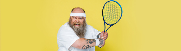 emocionado plus size homem com barba e raquete de tênis olhando para câmera isolada em amarelo, banner - Foto, Imagem