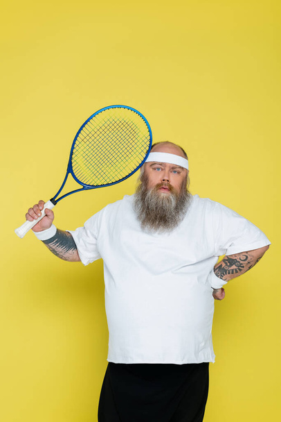 bearded plus size tennis player standing with hand on waist and looking at camera isolated on yellow - Zdjęcie, obraz