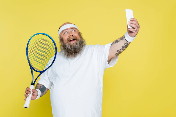 hombre feliz y regordete tomando selfie con raqueta de tenis aislado en amarillo - Foto, imagen