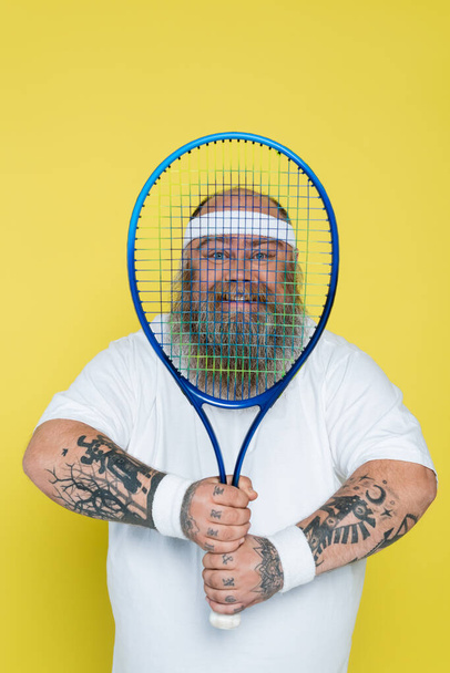 happy overweight man with beard looking at camera through tennis racquet isolated on yellow - Foto, Imagen