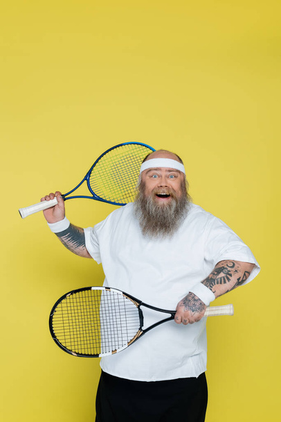 cheerful overweight tennis player with racquets looking at camera isolated on yellow - Foto, imagen