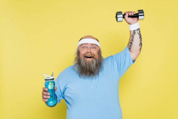 happy overweight man holding sports bottle while training with dumbbell isolated on yellow - Foto, imagen