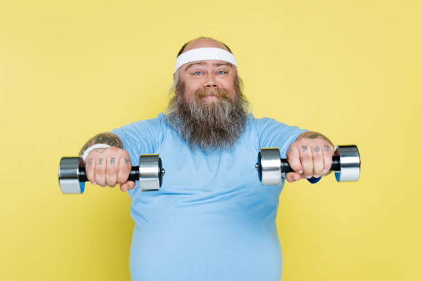 chubby and bearded man working out with dumbbells and smiling at camera isolated on yellow - Fotografie, Obrázek