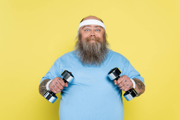 позитивный бородатый мужчина с избыточным весом, держащий гантели и глядя на камеру, изолированную на желтый - Фото, изображение