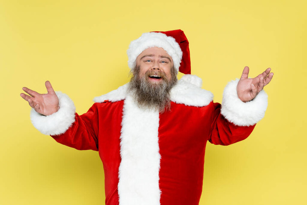 Радостный и пухлый Санта Клаус с приветственным жестом, изолированным на желтом - Фото, изображение