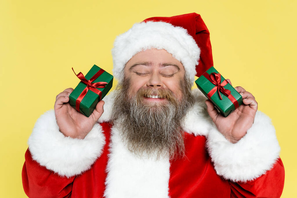 задоволений і пухкий Санта Клаус з закритими очима тримає подарункові коробки ізольовані на жовтому
 - Фото, зображення