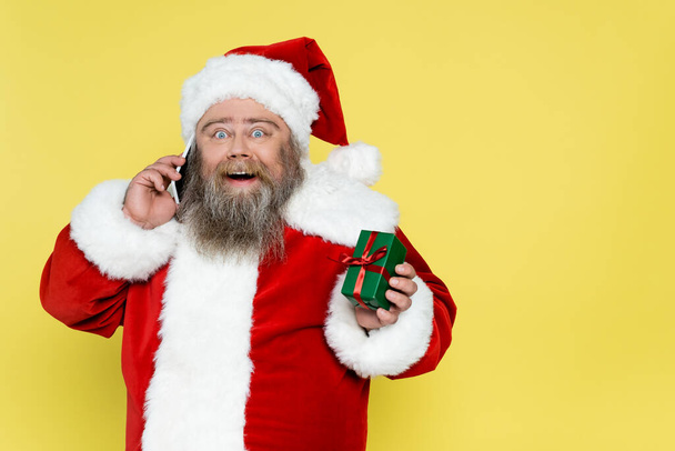 erstaunt plus größe weihnachtsmann zeigt geschenkbox, während sie auf smartphone isoliert auf gelb - Foto, Bild