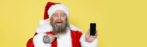 cheerful plus size santa claus pointing at mobile phone with blank screen isolated on yellow, banner - Photo, image
