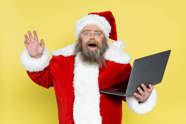 изумленный толстый Санта Клаус с ноутбуком размахивая рукой изолированы на желтый - Фото, изображение