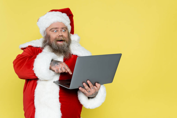 amazed overweight santa claus pointing at laptop isolated on yellow - Fotó, kép