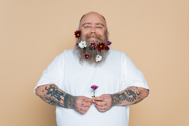 positive overweight man with decorated beard holding flower isolated on beige - Foto, imagen