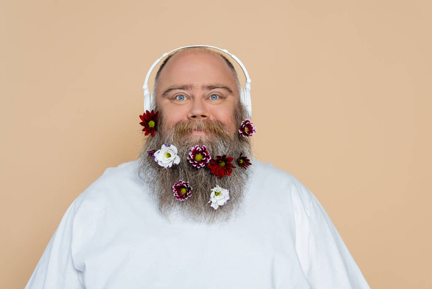 feliz hombre con sobrepeso con decoración floral en barba escuchando música en auriculares aislados en beige - Foto, imagen