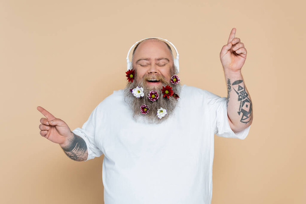 chubby and cheerful man with decorated beard listening music in headphones isolated on beige - Photo, Image
