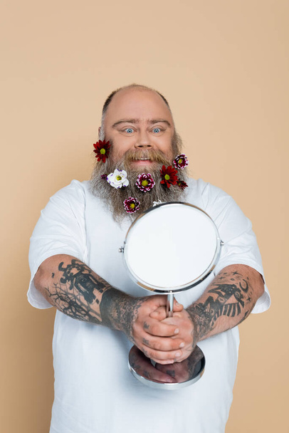 zadowolony plus size mężczyzna z kwiatowym wystrojem w brodzie patrząc w lustro odizolowane na beżu - Zdjęcie, obraz