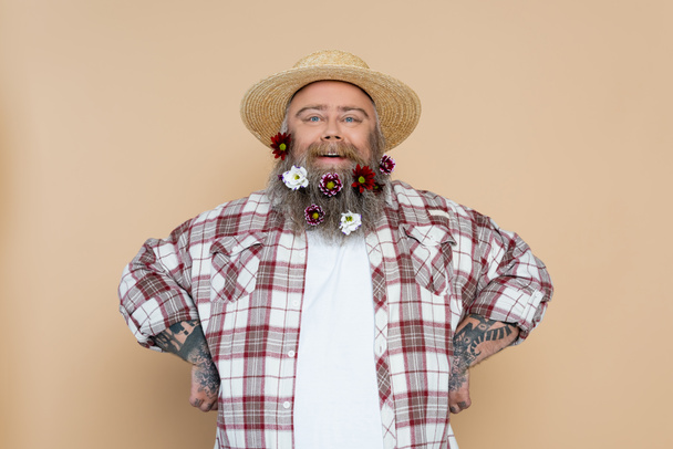 gelukkig overgewicht man in stro hoed en geruite shirt poseren met bloemen in baard geïsoleerd op beige - Foto, afbeelding