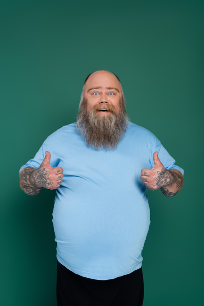 γενειοφόρος υπέρβαρος άντρας με μπλε μπλουζάκι χαμογελώντας στην κάμερα και δείχνοντας τους αντίχειρες επάνω απομονωμένους στο πράσινο - Φωτογραφία, εικόνα