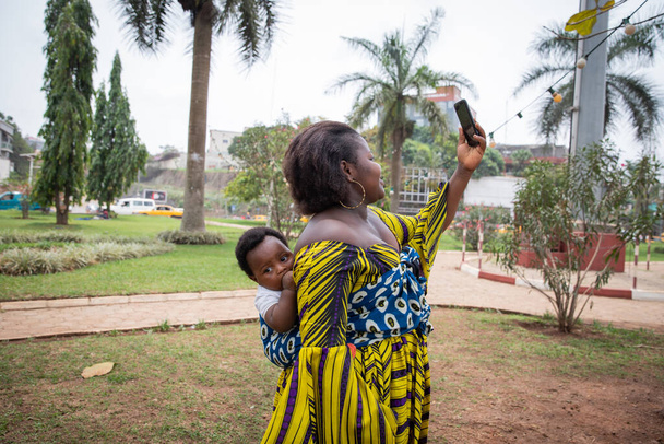 African mother with her son held on her back takes a selfie with her smartphone - Foto, Imagen