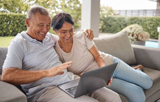 Senior couple making a video call on a laptop at home. Two happy people browsing the internet. Elderly mixed race man and woman relaxing on the sofa, hugging and laughing while using modern computer. - Fotoğraf, Görsel