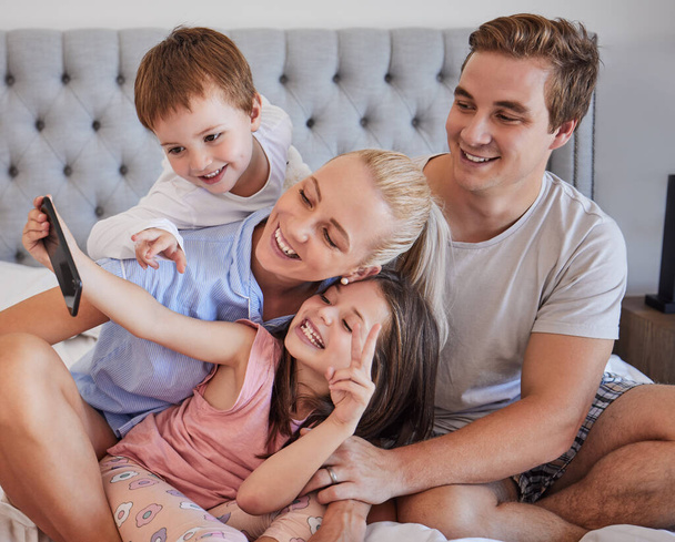Happy young caucasian family sitting together on bed and taking a selfie or doing video call together in the morning. Carefree little girl holding mobile phone while recording video of special moment - Zdjęcie, obraz