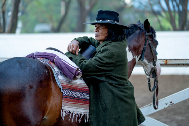 Asiatischer alter Mann im Cowboykostüm steht und lehnt neben einem dunkelbraunen Pferd und schaut nach links und bleibt in der Nähe des Stalls, um das Pferd zu trainieren. - Foto, Bild