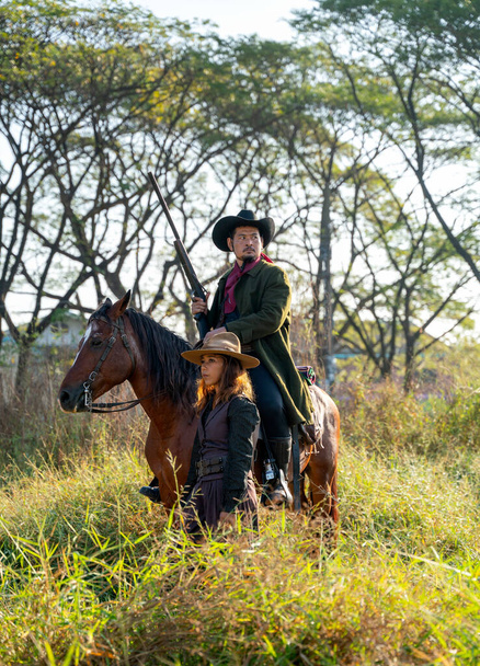 Junge Asiatin im Cowgirl-Kostüm steht vor Cowboy auf Pferd mit haltender Waffe und sieht aus wie Schutz und Geborgenheit im Feld - Foto, Bild