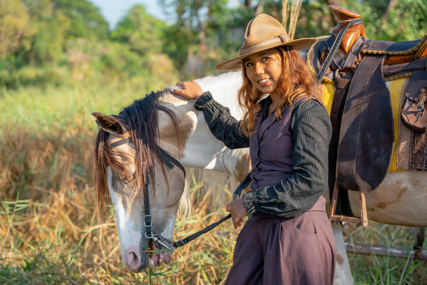 Bastante joven mujer asiática con traje de vaquero sonrisa y de pie delante de caballo blanco y también mirar a la cámara en el campo. - Foto, imagen