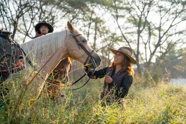 Csinos fiatal ázsiai nő cowboy jelmez mosollyal, és álljon előtt fehér ló, és maradjon közel más cowboy férfi a területen a falu közelében. - Fotó, kép