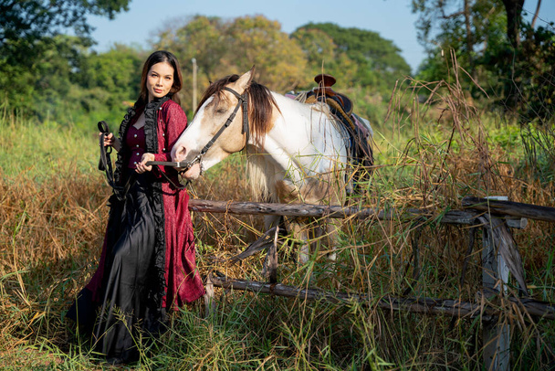 Kaunis nuori aasialainen nainen seistä ja antaa aidan lähellä valkoinen hevonen myös katsoa kameran alalla. - Valokuva, kuva
