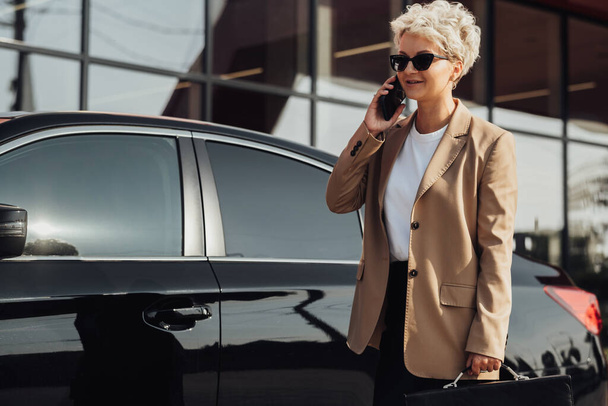 Elegante Geschäftsfrau mit Aktentasche in der Hand telefoniert in der Nähe des Luxusautos - Foto, Bild